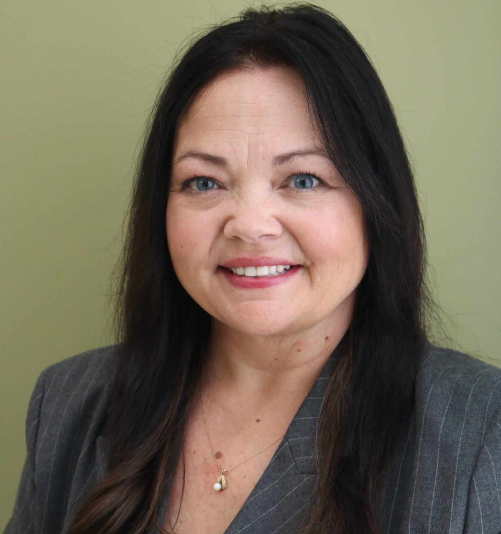 Shannon Olsson, US Product Manager, is a white woman with long dark hair wearing a grey pinstripe blazer and a gold necklace, smiling in front of a light green background