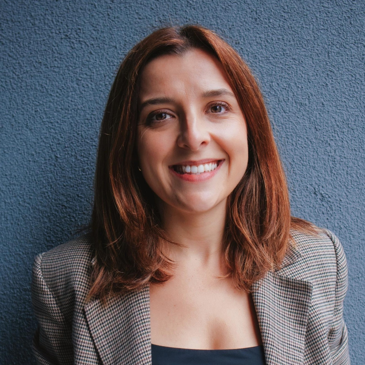 Square portrait of Sarita Gianesini smiling, with long dark hair, wearing a beige blazer against a neutral background