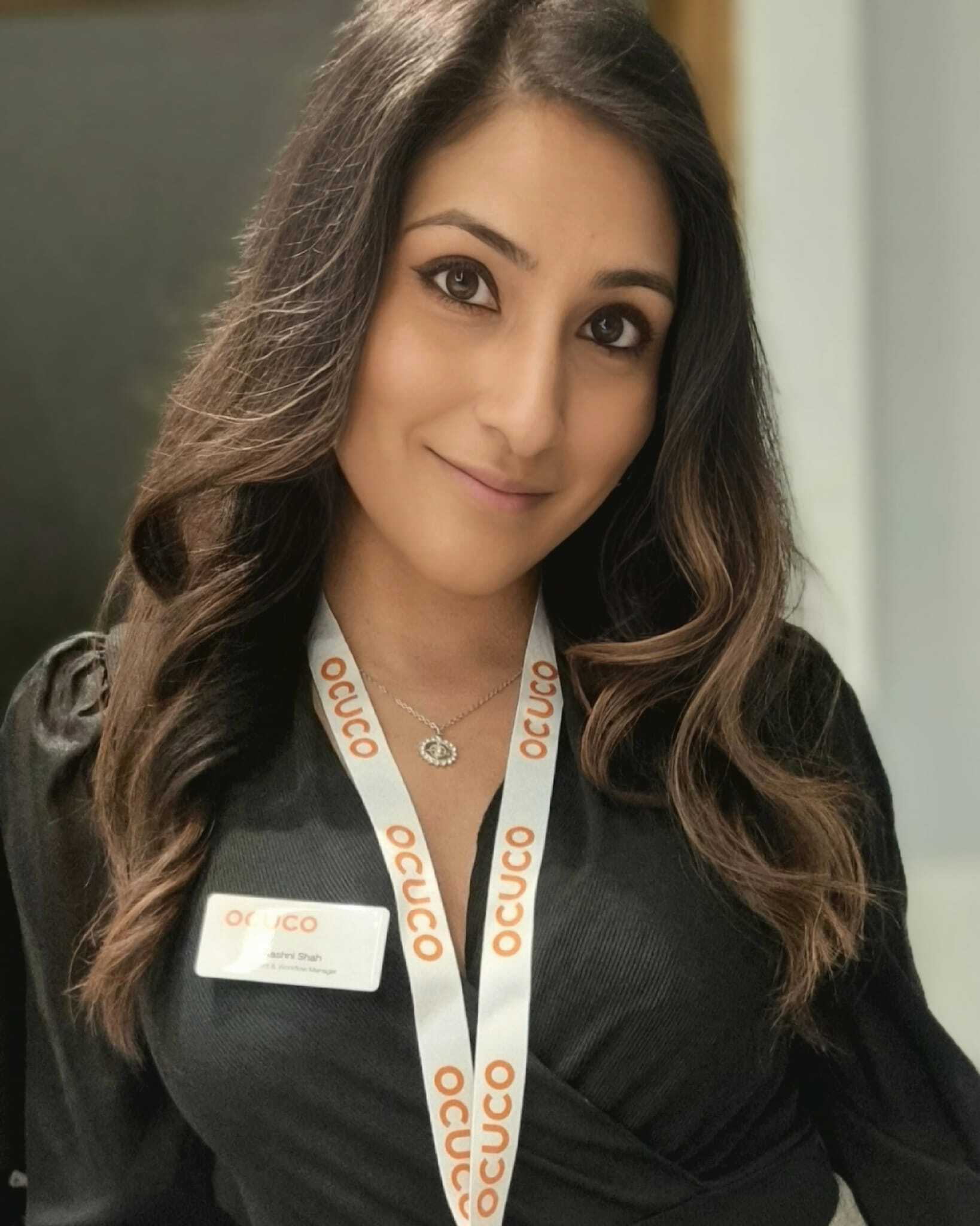 Aashni Shah, with long wavy brown hair, wearing a black wrap-style blouse and a silver necklace, smiles at the camera. She has a white Ocuco lanyard with orange lettering and a visible name badge, standing in an indoor professional setting with a blurred background.