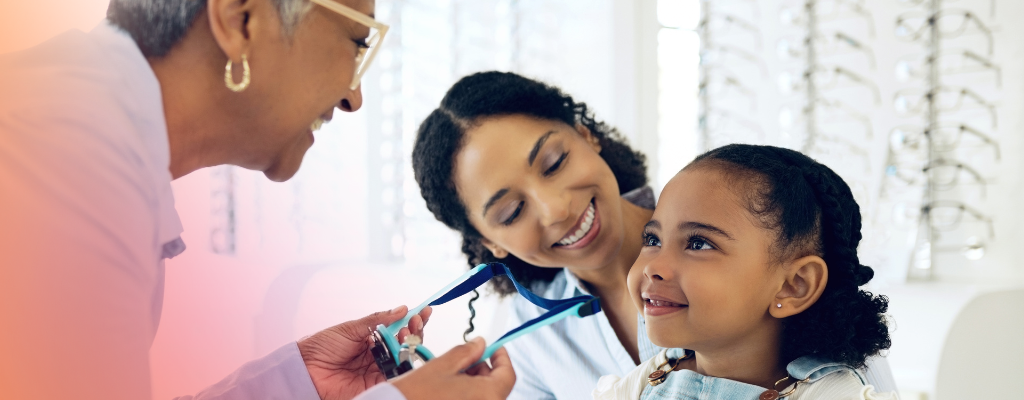 Optician showing a pair of glasses to a smiling young girl during an eyecare appointment, with the child’s mother watching beside her in an optical practice.