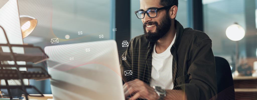 A man wearing glasses sits at a desk smiling while working on a laptop, with small illustrated email icons floating around him to represent email marketing for opticians.